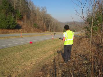 A researcher in a yellow safety vest stands on the edge of a four lane road