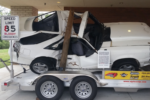 crushed white car on truck trailer bed next to bent 85 mile per hour sign