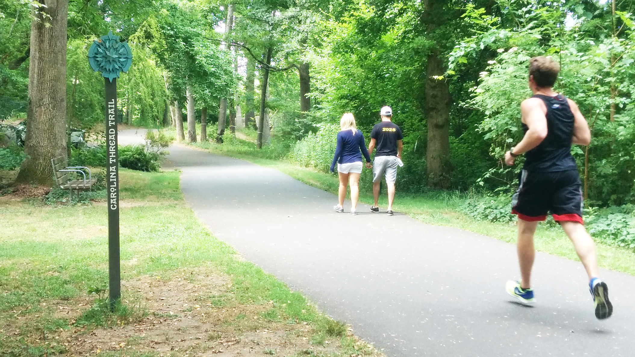 jogger and walkers pass the "Carolina Thread Trail" marker of a tree lined greenway