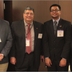 Four men in suits with name tags pose in front of a conference room wall, including Nagui and Shams
