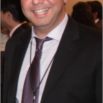 Behzad smiling in a suit at a conference, with other business professionals obscured but visible in the background
