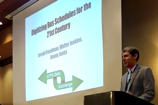 ITRE research assistant Jonah Freedman, presenting at the NC Public Transportation Association conference, with a projector presentation showing that reads "Digitizing bus schedules for the 21st century. Jonah Freedman, Walter Jenkins, Jimmy Jones"