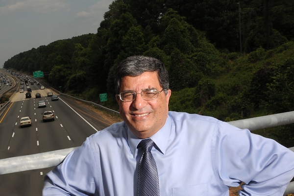 A man in a dress shirt (Nagui Rouphail) poses against the side guard of a highway I-40 overpass, with cars passing underneath