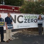 ITRE researchers stand before an NC Vision Zero banner and a Morrisville firetruck, holding signs that read "Take The Pledge, ncvisionzero.org" and "save lives through safer roads"