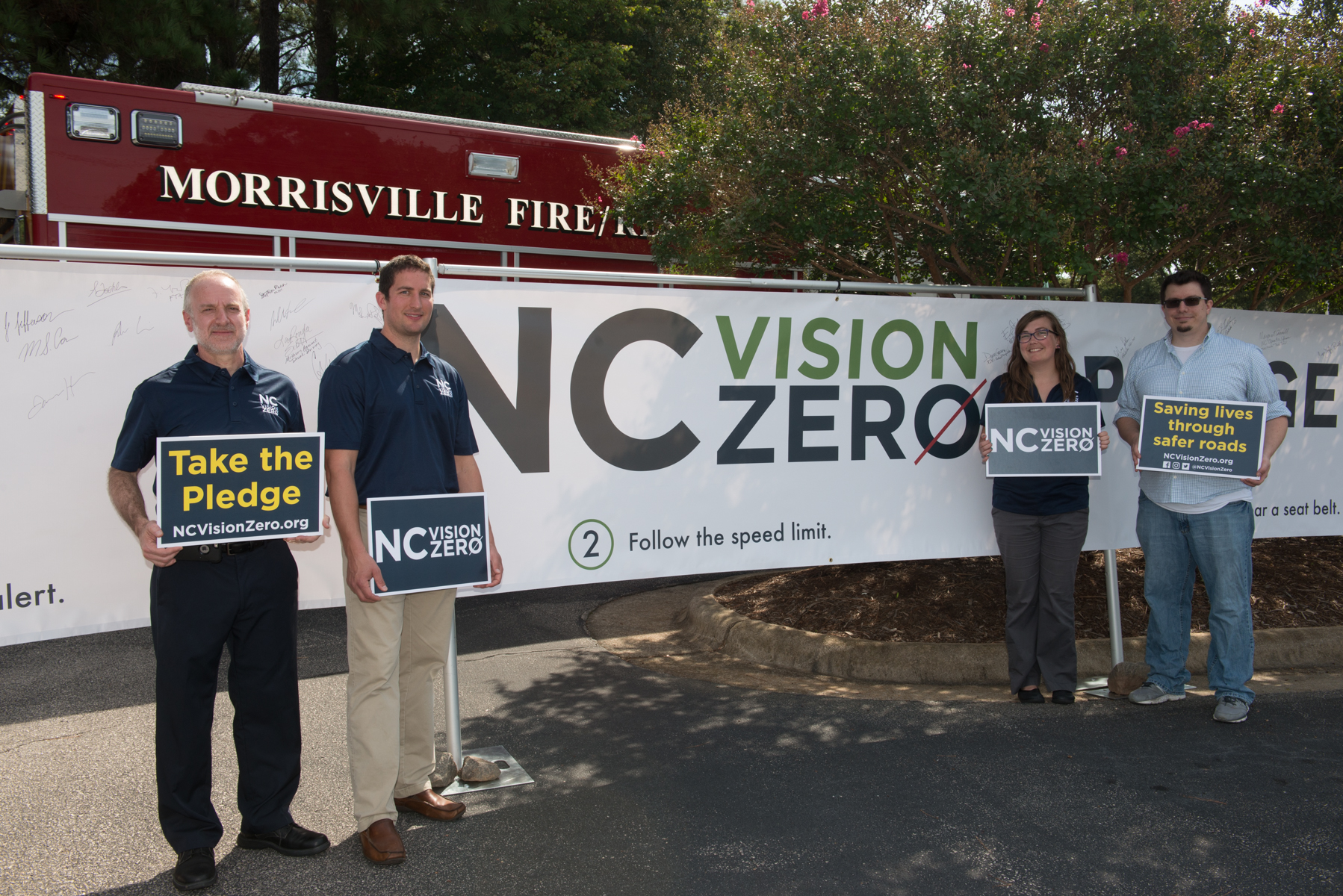 ITRE researchers stand before an NC Vision Zero banner and a Morrisville firetruck, holding signs that read "Take The Pledge, ncvisionzero.org" and "save lives through safer roads"