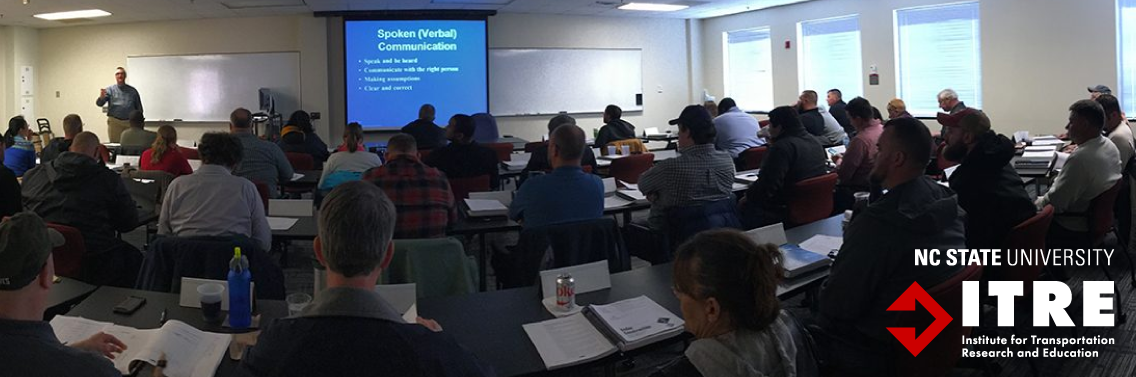 About 30 people sitting in a classroom, with a presenter at the front. A projector screen reads "Spoken (Verbal) Communication."