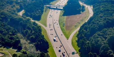 Aerial view of a highway with light traffic. Dense trees dominate the adjacent landscape.