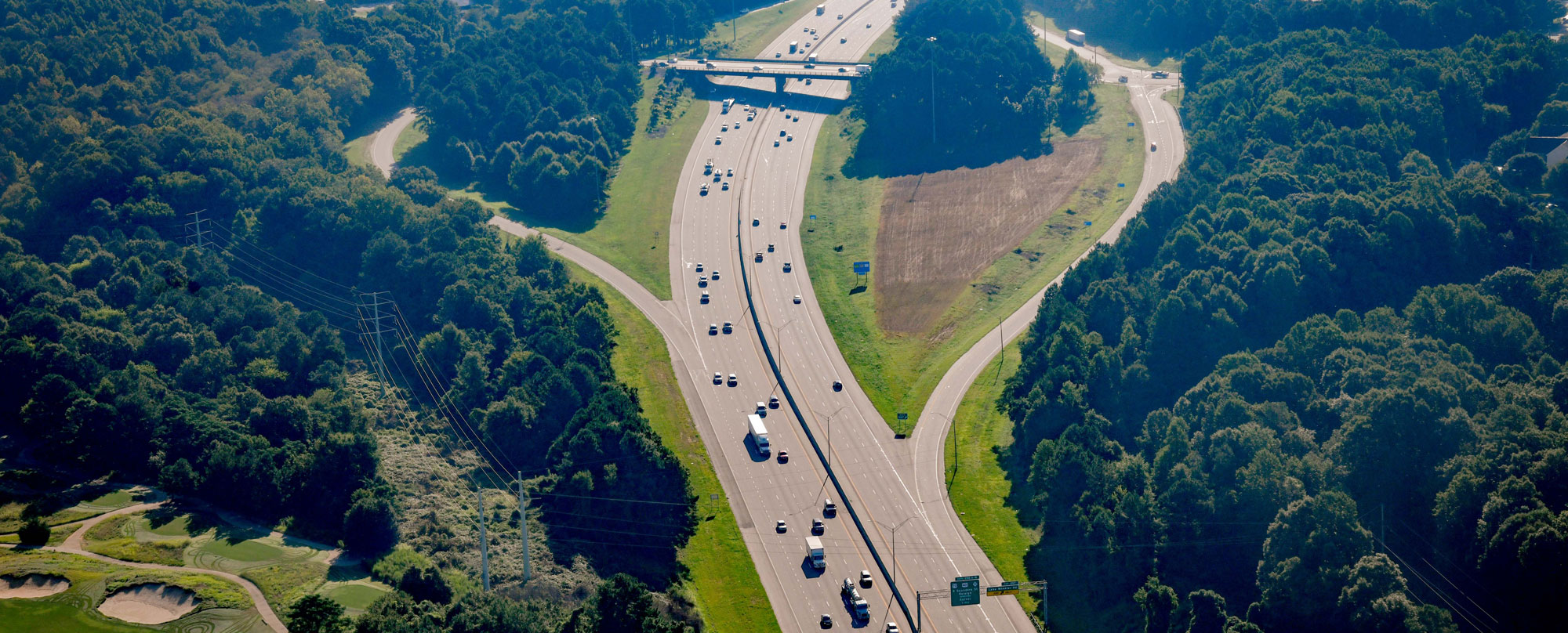 Aerial view of a highway with light traffic. Dense trees dominate the adjacent landscape.