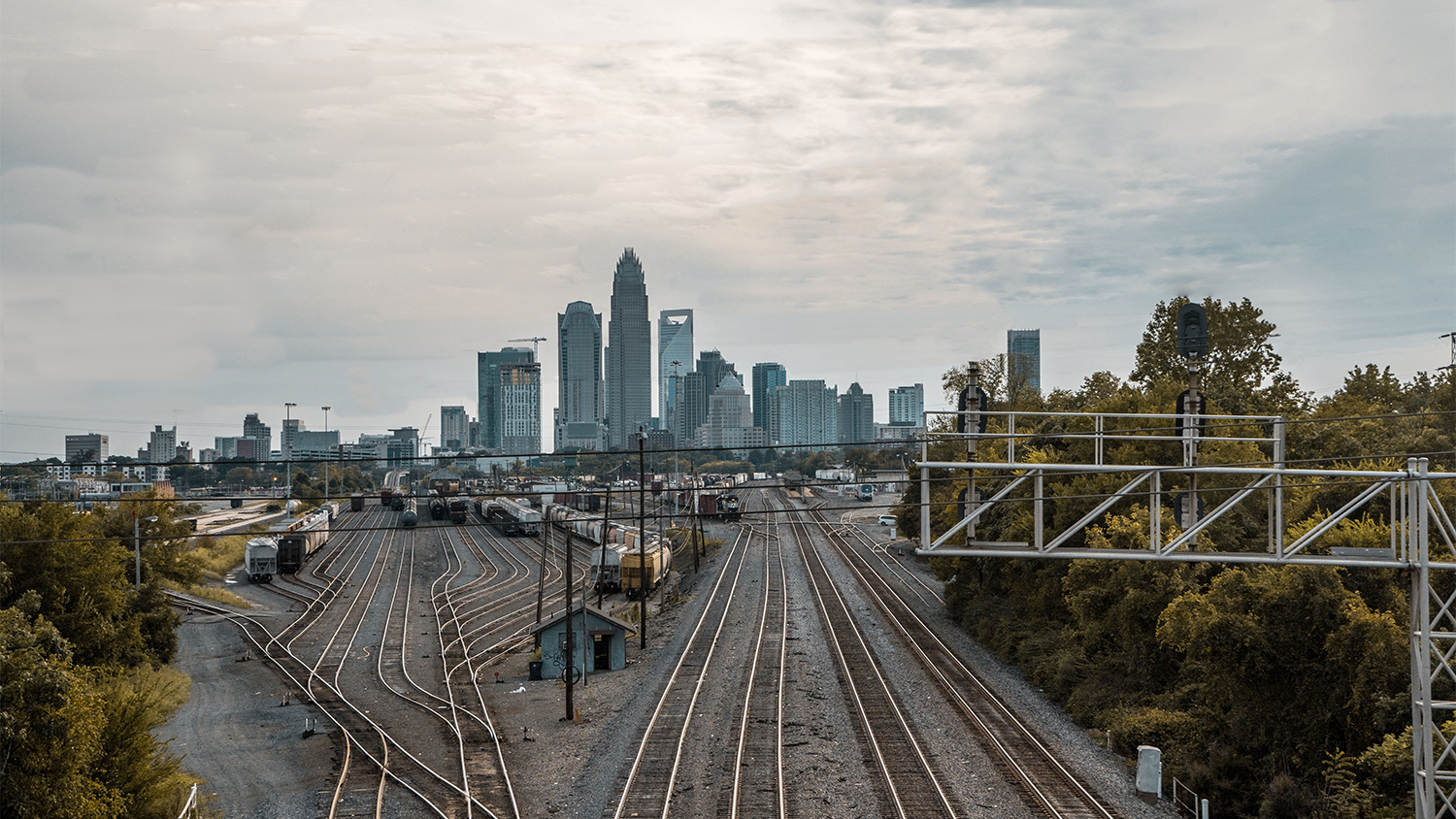 The city skyline of Charlotte looms over a rail yard with multiple train cars stored on several tracks