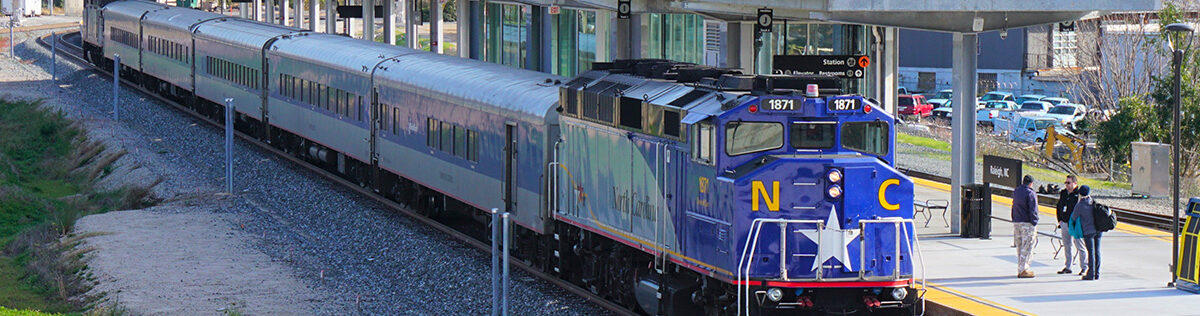 A passenger train sits at a Raleigh station platform on a sunny day. The engine design is inspired by the North Carolina state flag, with blue paint and yellow 