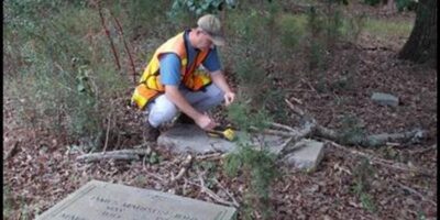 A person with light skin wearing a high-visibility vest and baseball cap squats next to a gravestone in a wooded area. Another gravestone with visible engravings is visible in the foreground.