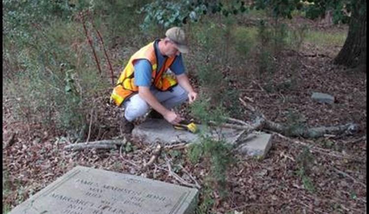 A person with light skin wearing a high-visibility vest and baseball cap squats next to a gravestone in a wooded area. Another gravestone with visible engravings is visible in the foreground.