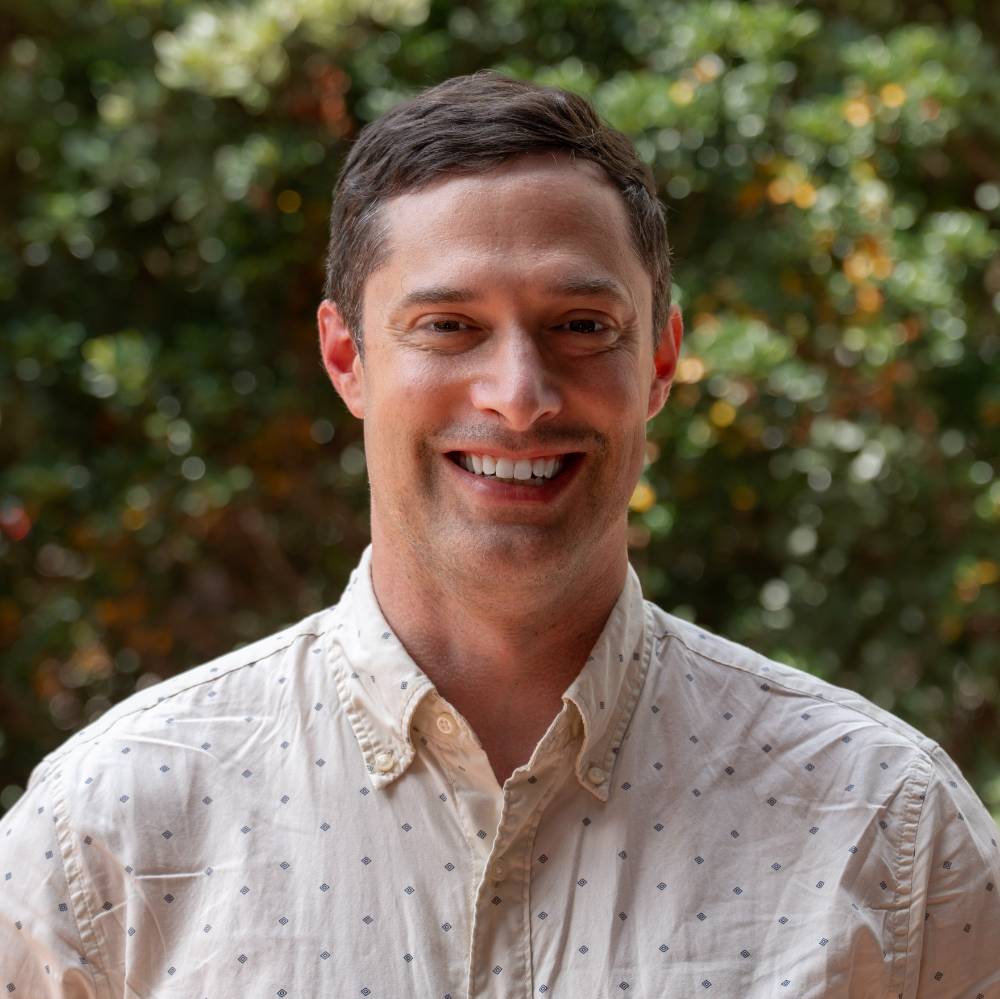A smiling person with short brown hair, wearing a light-colored button-up shirt with a repeating pattern. They are standing outside in front of blurred foliage.