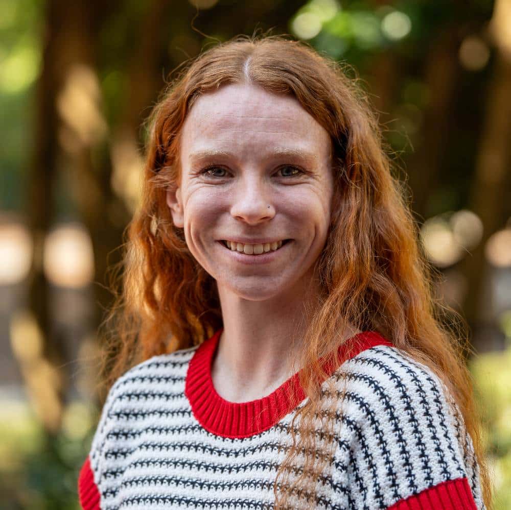 A smiling person with long, wavy reddish-brown hair, wearing a black, white, and red sweater, standing outside in front of a blurred foliage background.