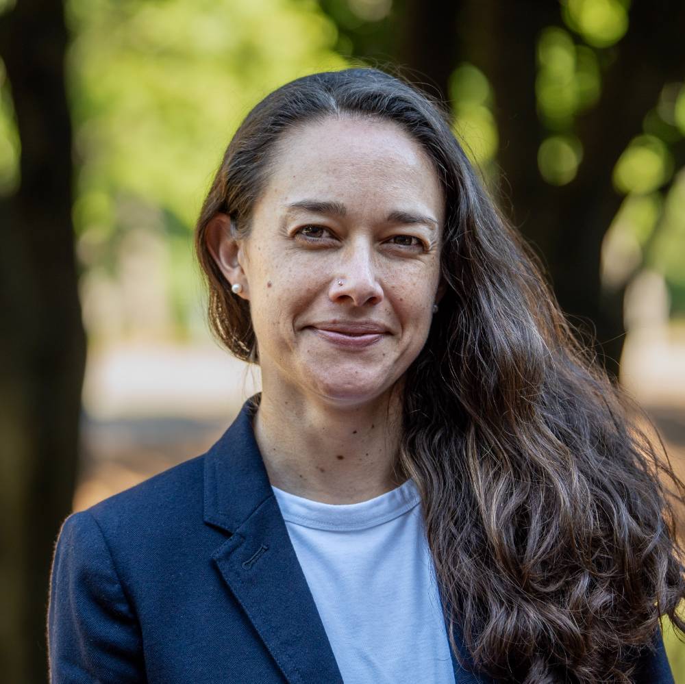 A person with a light skin tone and long, wavy, dark hair draped over one shoulder. They are wearing a dark blue blazer over a white shirt, a small nose stud, and pearl earrings, set against a blurred background of green foliage.