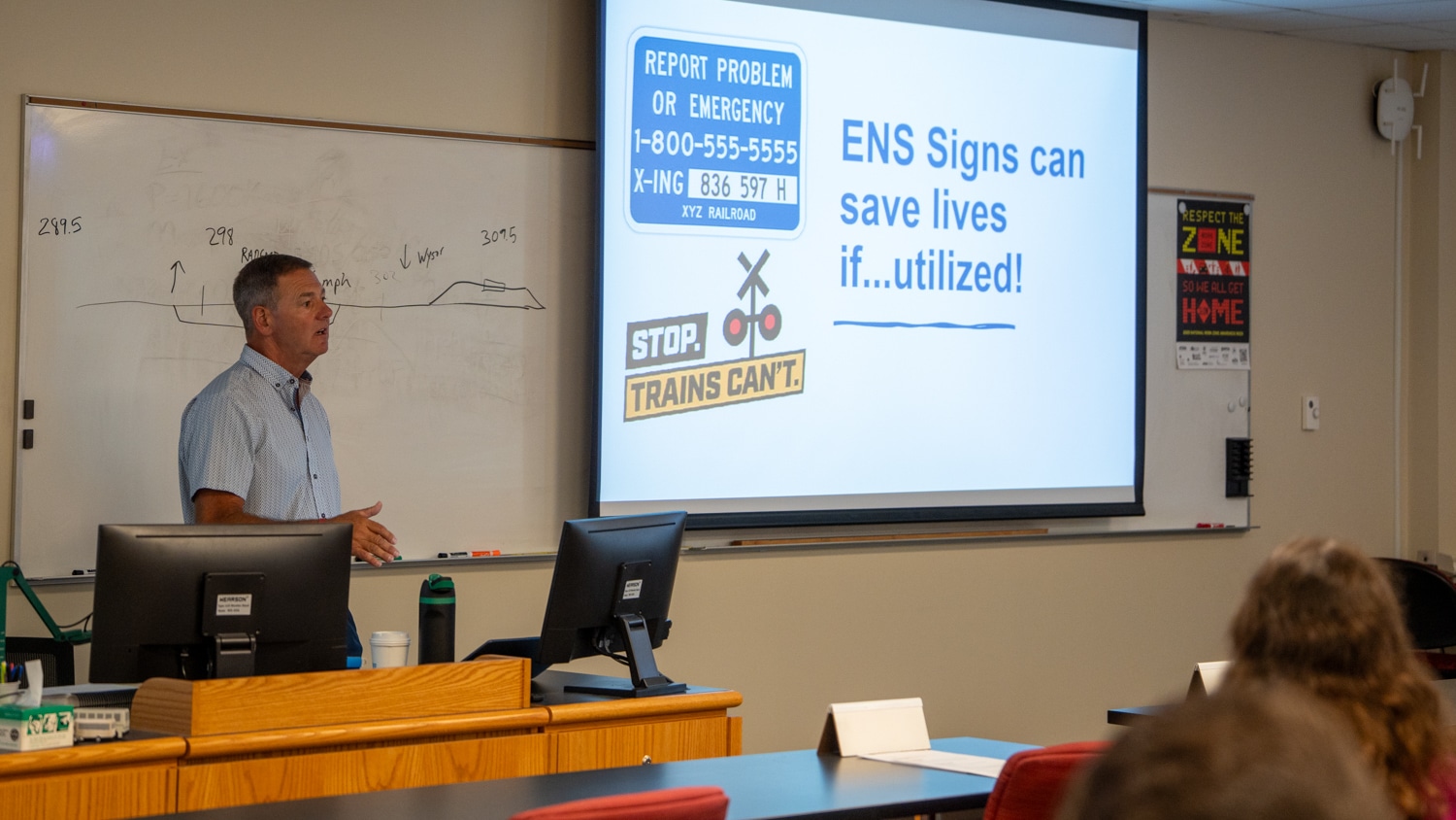 A presenter in a classroom gives a lecture on railroad safety next to a slide display about ENS signs.